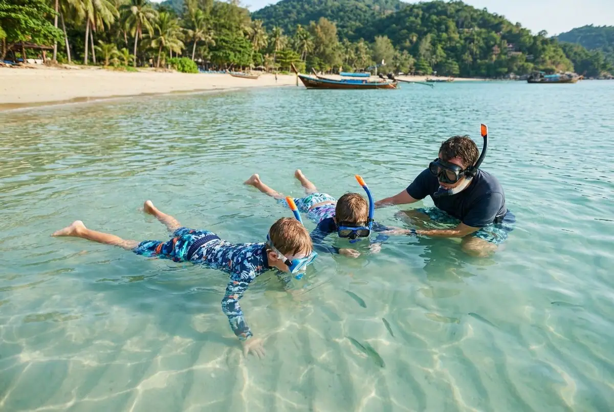 Two kids in masks and rash guards snorkeling in calm, clear shallow water over sand, a parent beside them